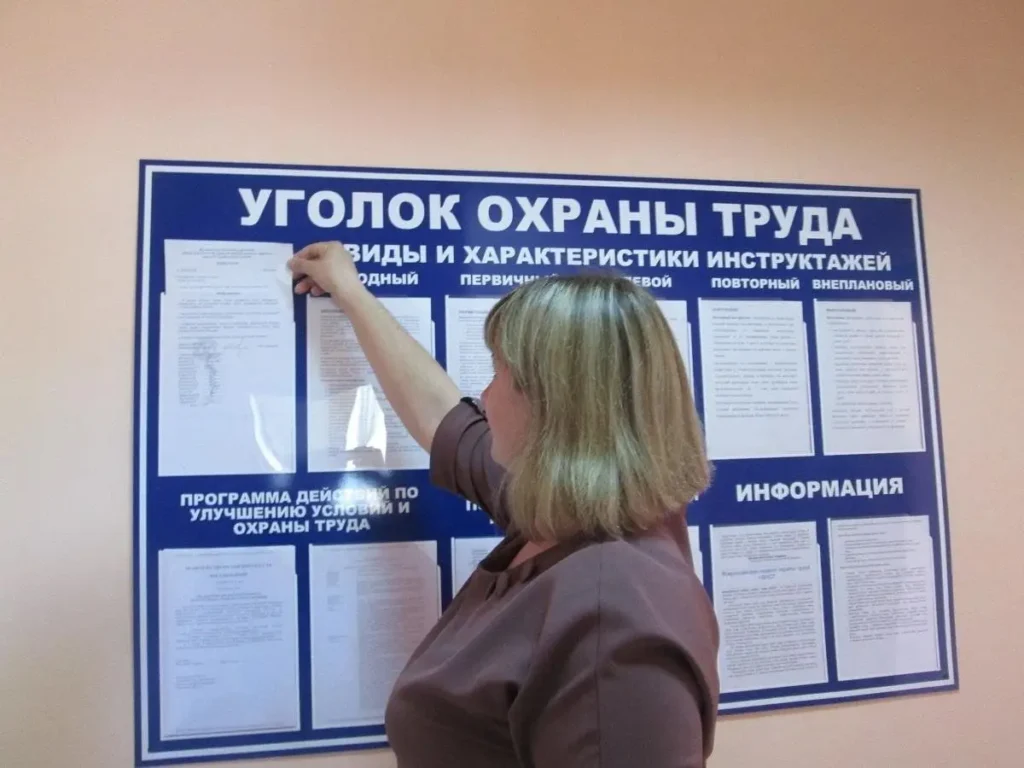 Женщина изучает информационный стенд "Уголок охраны труда" с документами о видах инструктажей и программах улучшения условий труда